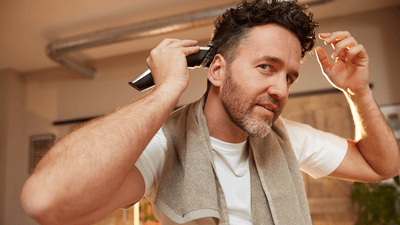 A man is using a black and grey Philips hair clipper to shave and style the right side of his head.