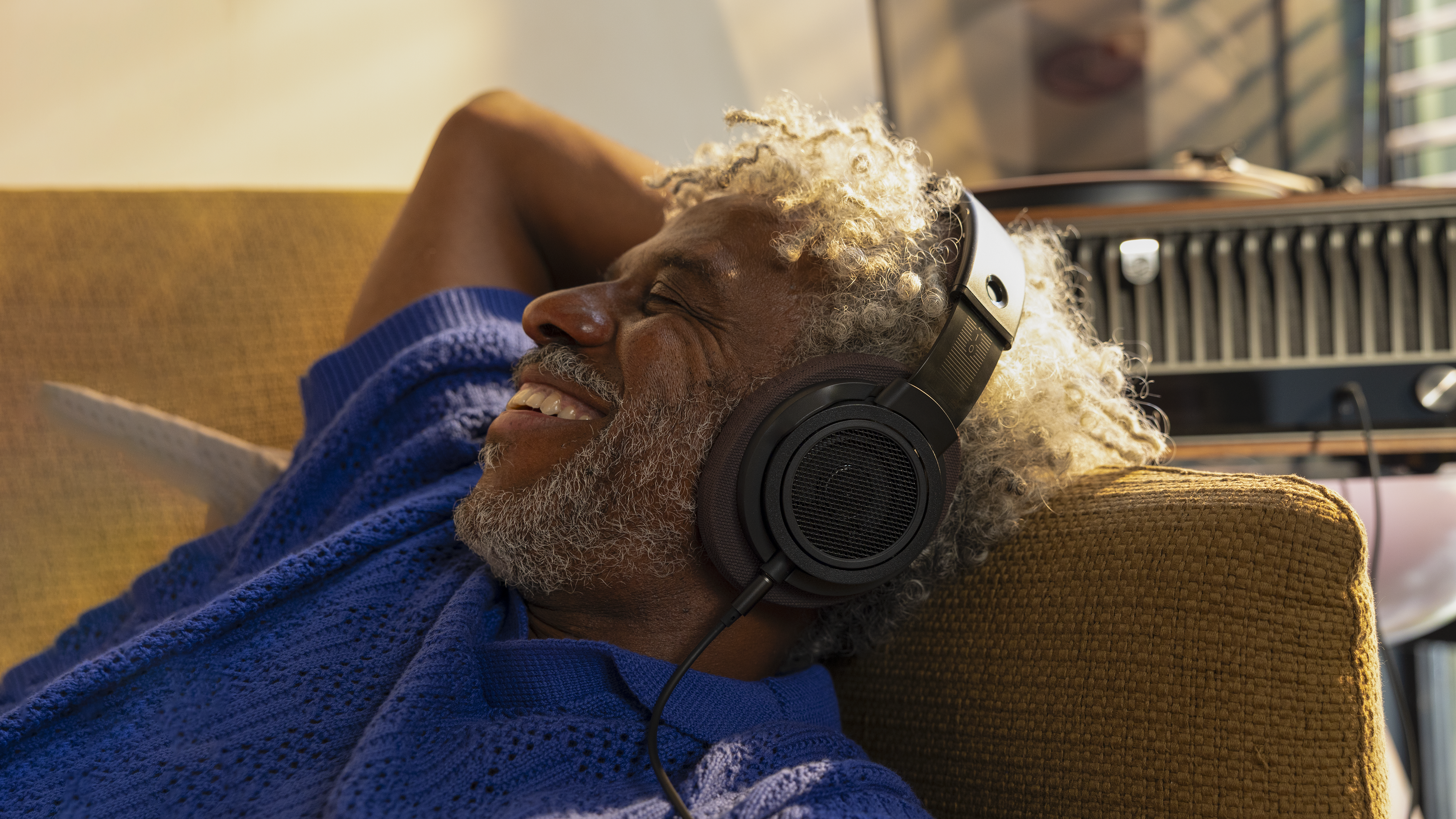 Starejši moški s kodrastimi sivimi lasmi se sprošča na zofi v udobnem domačem okolju, miži in se smehlja, medtem ko posluša glasbo prek naušesnih slušalk Philips.