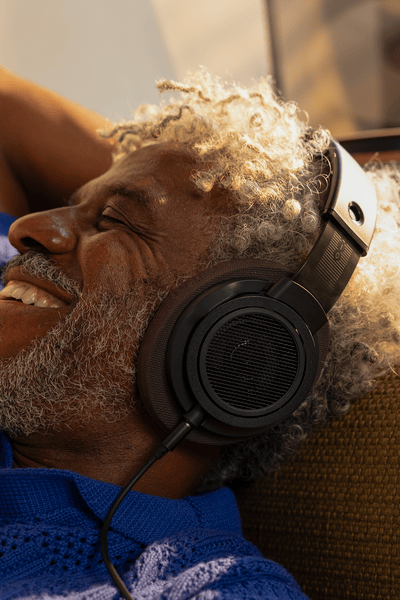 Starejši moški s kodrastimi sivimi lasmi se sprošča na zofi v udobnem domačem okolju, miži in se smehlja, medtem ko posluša glasbo prek naušesnih slušalk Philips.