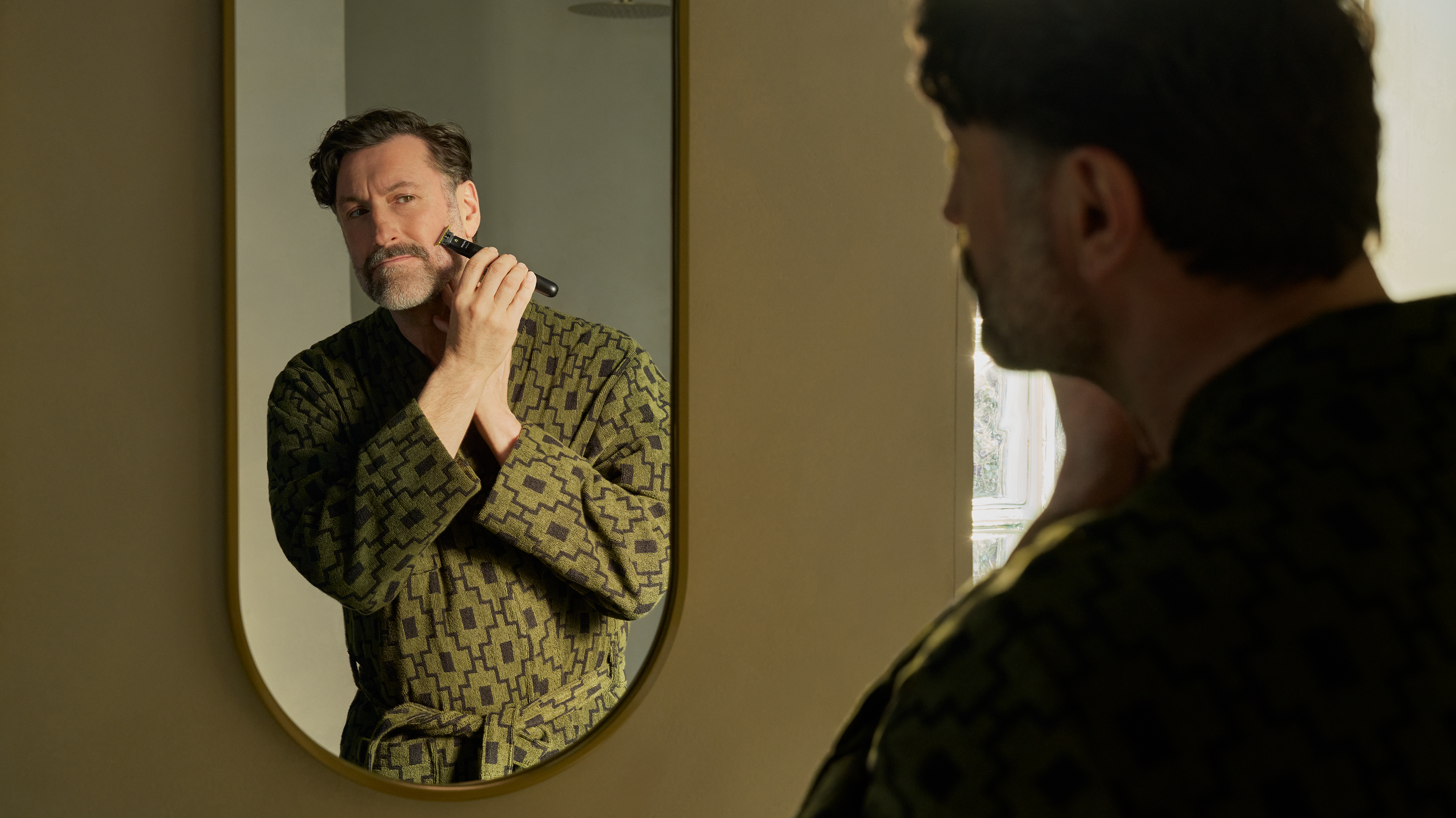 A man in a green patterned bathrobe trims his moustache with a Philips OneBlade.
