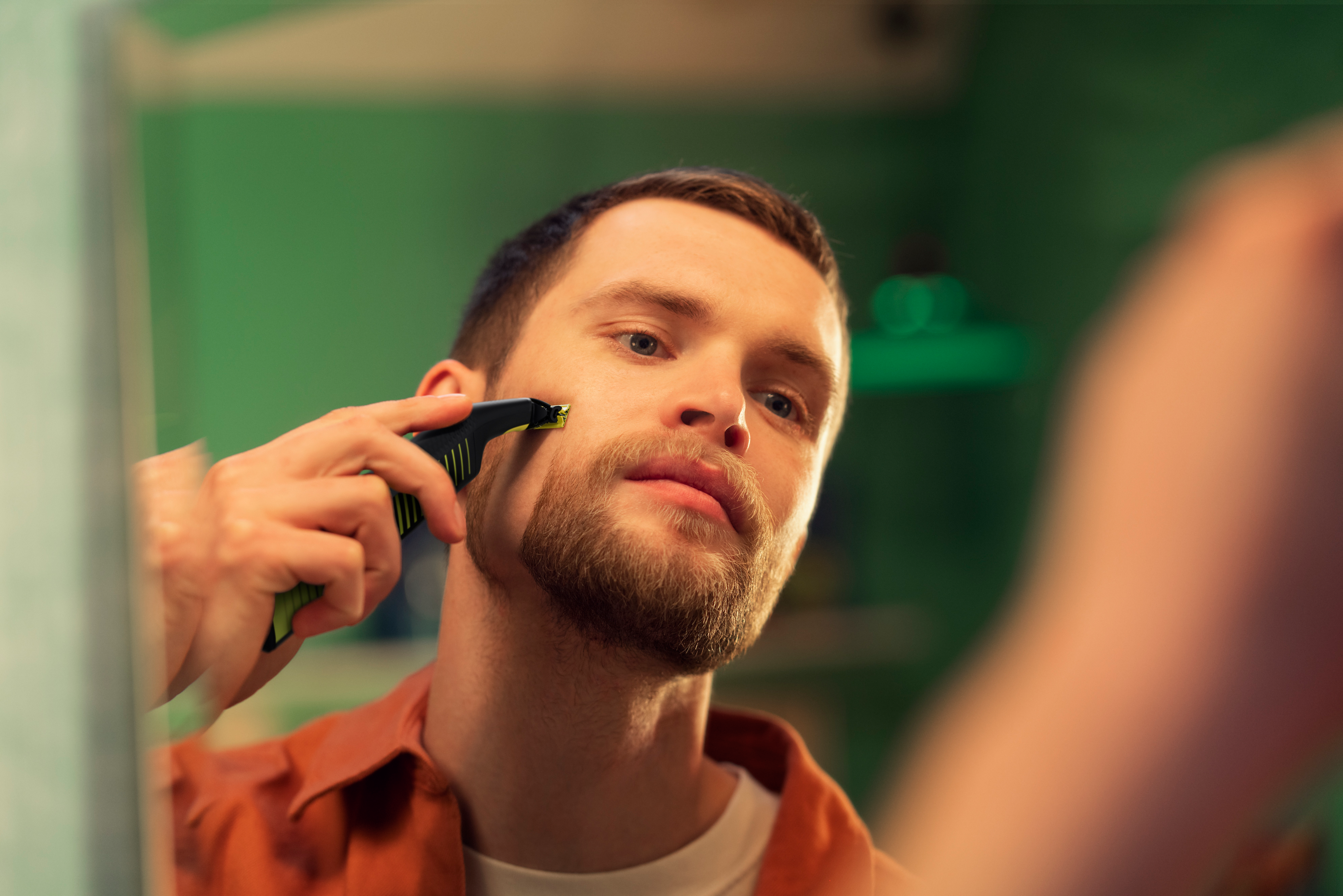 Ein Mann in einem orangefarbenen Hemd rasiert seinen Vollbart mit dem Philips OneBlade in einem Badezimmerspiegel.