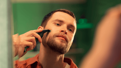 Ein Mann in einem orangefarbenen Hemd rasiert seinen Vollbart mit dem Philips OneBlade in einem Badezimmerspiegel.