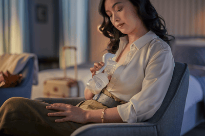 A smartly dressed woman expresses milk using a Philips Avent electric breast pump in a dimly lit room.