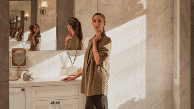 A woman with dark hair stands in her dressing room with her back to a mirror and one hand resting gently on her face.