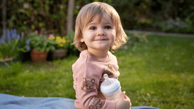 Ein blondes Kleinkind strahlt in die Kamera, während es eine Philips Avent Babyflasche in der Hand hält.