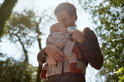 A man wearing a baby carrier feeds his infant with a Philips Avent baby bottle in an outdoor setting.