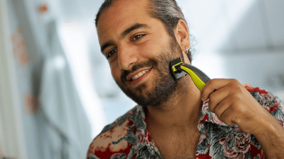 Un hombre con una camisa estampada sonríe mientras sujeta una cuchilla Philips OneBlade contra su espesa barba.