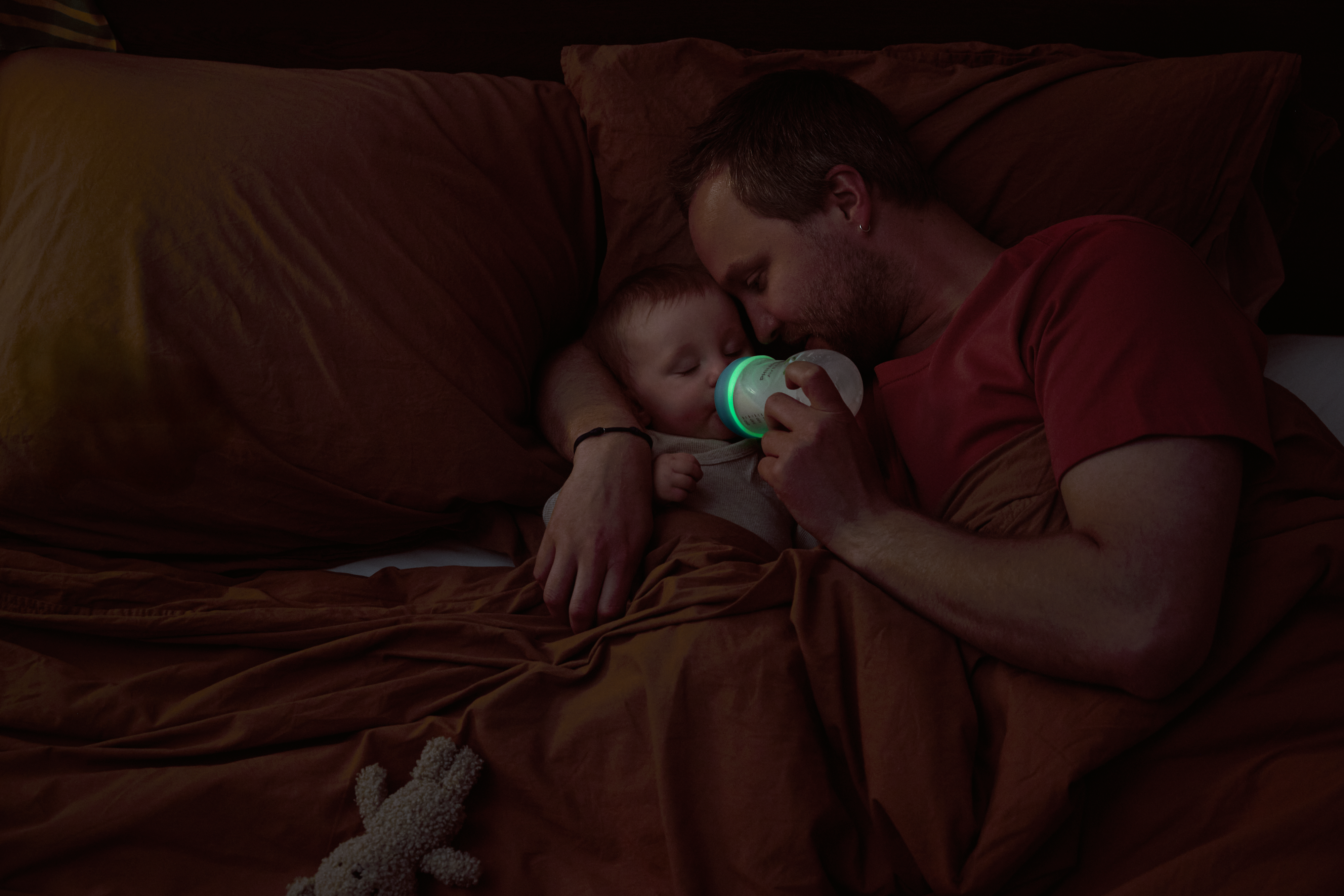 A man lies in bed with his baby, arm wrapped around them while feeding with a Philips Natural Response Nighttime bottle.