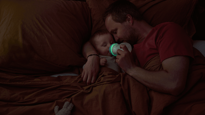 A man lies in bed with his baby, arm wrapped around them while feeding with a Philips Natural Response Nighttime bottle.