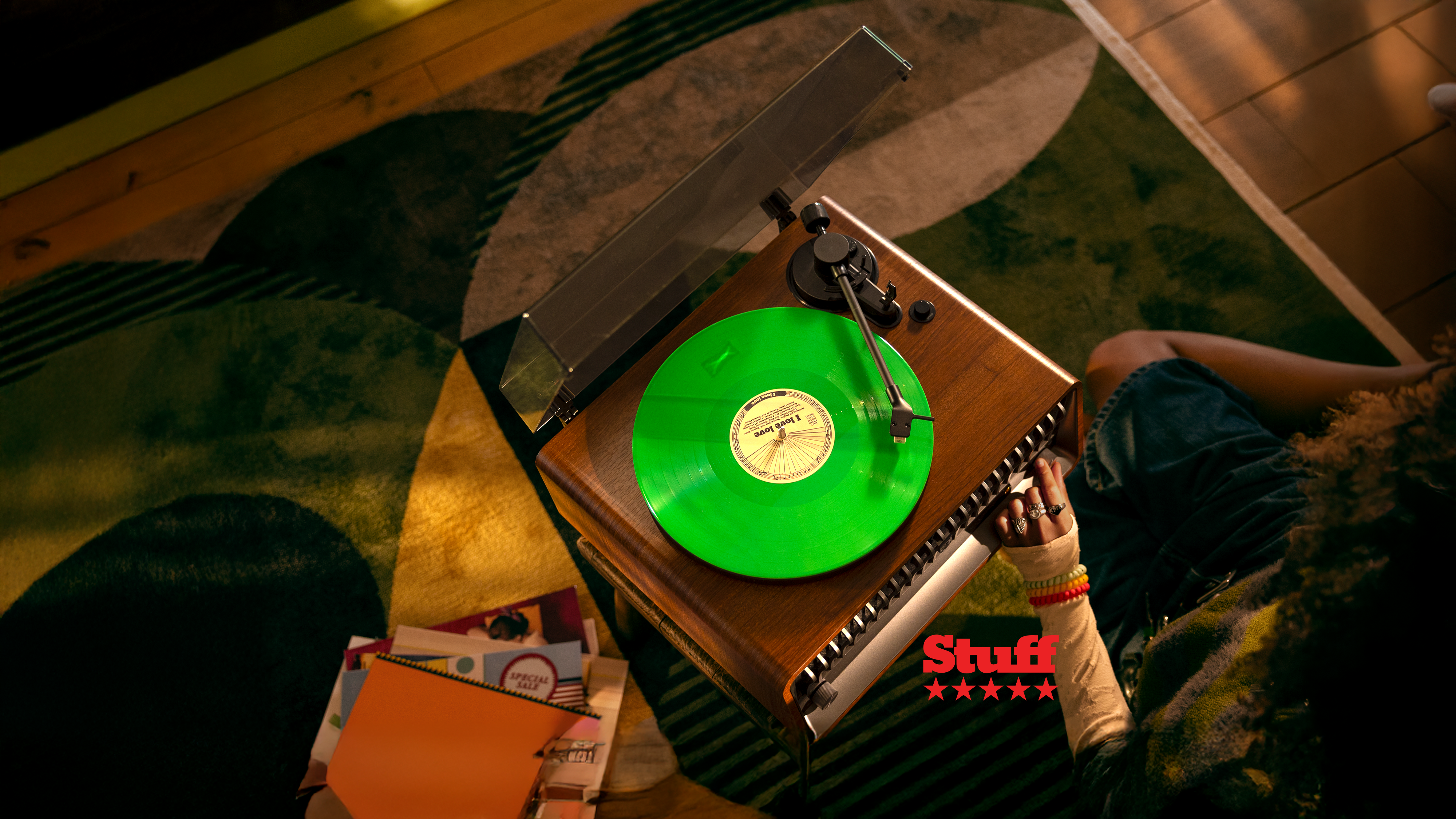 A top-down view of a bright green vinyl record playing on a wooden turntable in a cozy home setting, with a person relaxing nearby and notebooks scattered on the table, highlighting a Philips sound experience.