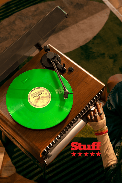 A top-down view of a bright green vinyl record playing on a wooden turntable in a cozy home setting, with a person relaxing nearby and notebooks scattered on the table, highlighting a Philips sound experience.
