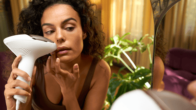 A young woman with dark curly hair leans towards a mirror to remove facial hair with a grey Philips Lumea device