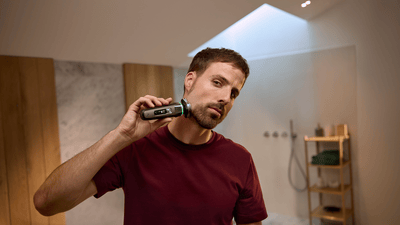 A man looking directly at the camera is shaving his chin area using a blue Philips shaver.