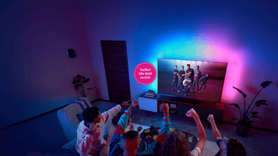 Philips Ambilight TV mit FC-Barcelona-Spielern auf dem Bildschirm, während eine Gruppe von Personen auf einem Sofa in einem abgedunkelten Wohnzimmer mit farbigem Ambientelicht zusieht und jubelt.