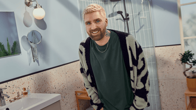 A man with blond hair and a dark beard is wearing a cardigan while standing in a bathroom smiling at the camera.