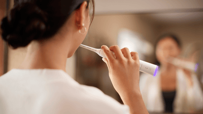 A woman with dark hair, wearing a white robe, is shown from behind brushing her teeth to prevent bad breath in pregnancy.