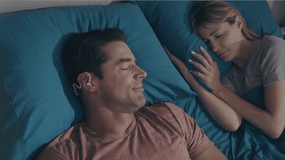 A man and a woman are sleeping in bed with the man wearing sleep headphones.