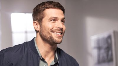 A man with a fashionable beard and a smart suit smiles as he looks off into the distance.