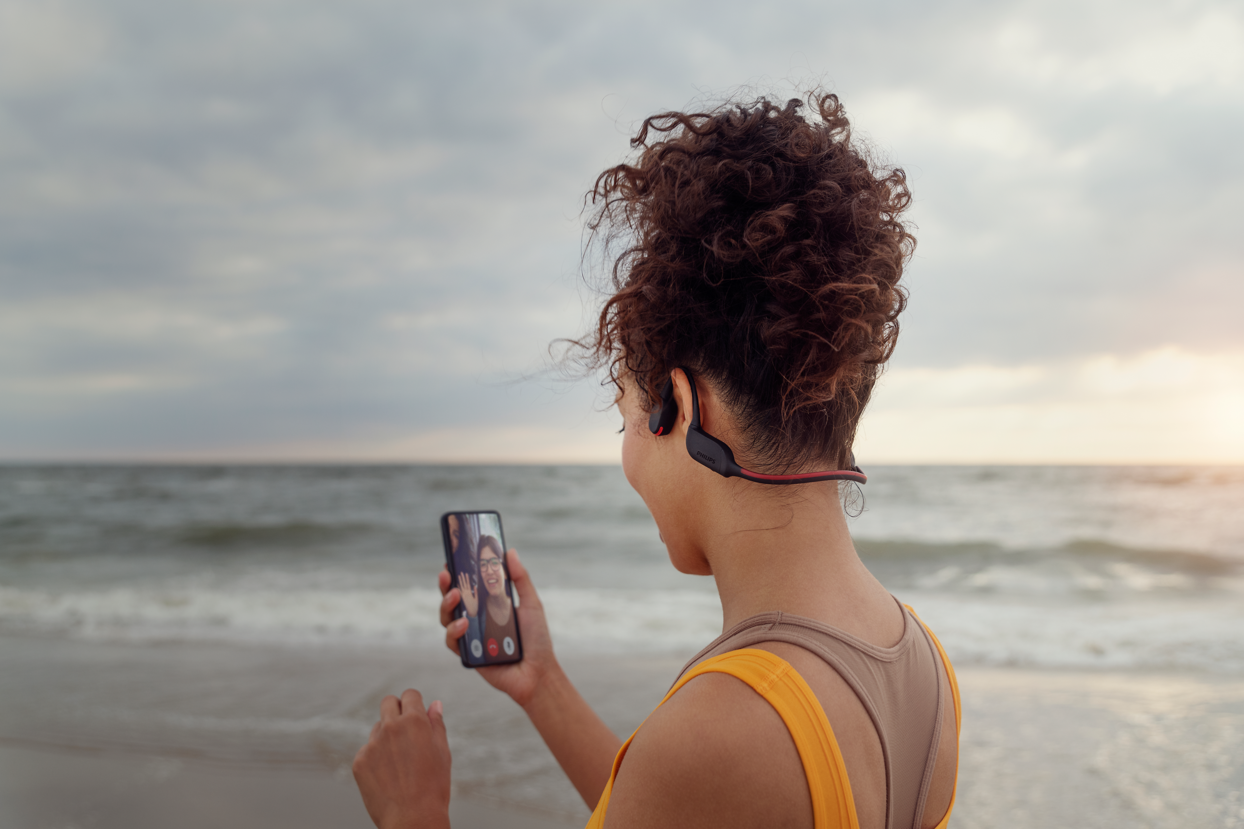 Športovec telefonujúci na pláži prostredníctvom použítia slúchadiel s kostným vedením zvuku