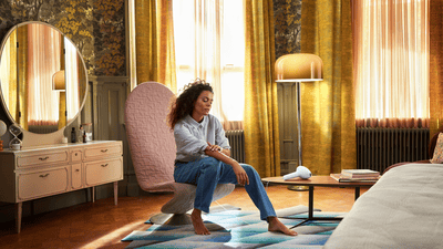 A woman wearing jeans and a button-down, sitting on a chair in her bedroom, looking down at her legs.