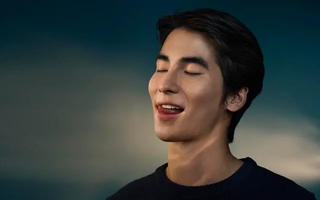 A young man smiles as he licks his white teeth against a dark gradient background.
