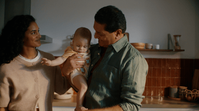 A couple in a kitchen holding their baby, who’s just been woken up to get fed.