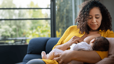 A woman wearing a bright yellow t-shirt breastfeeds her young baby while sitting on a dark blue sofa.