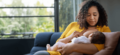 A woman wearing a bright yellow t-shirt breastfeeds her young baby while sitting on a dark blue sofa.