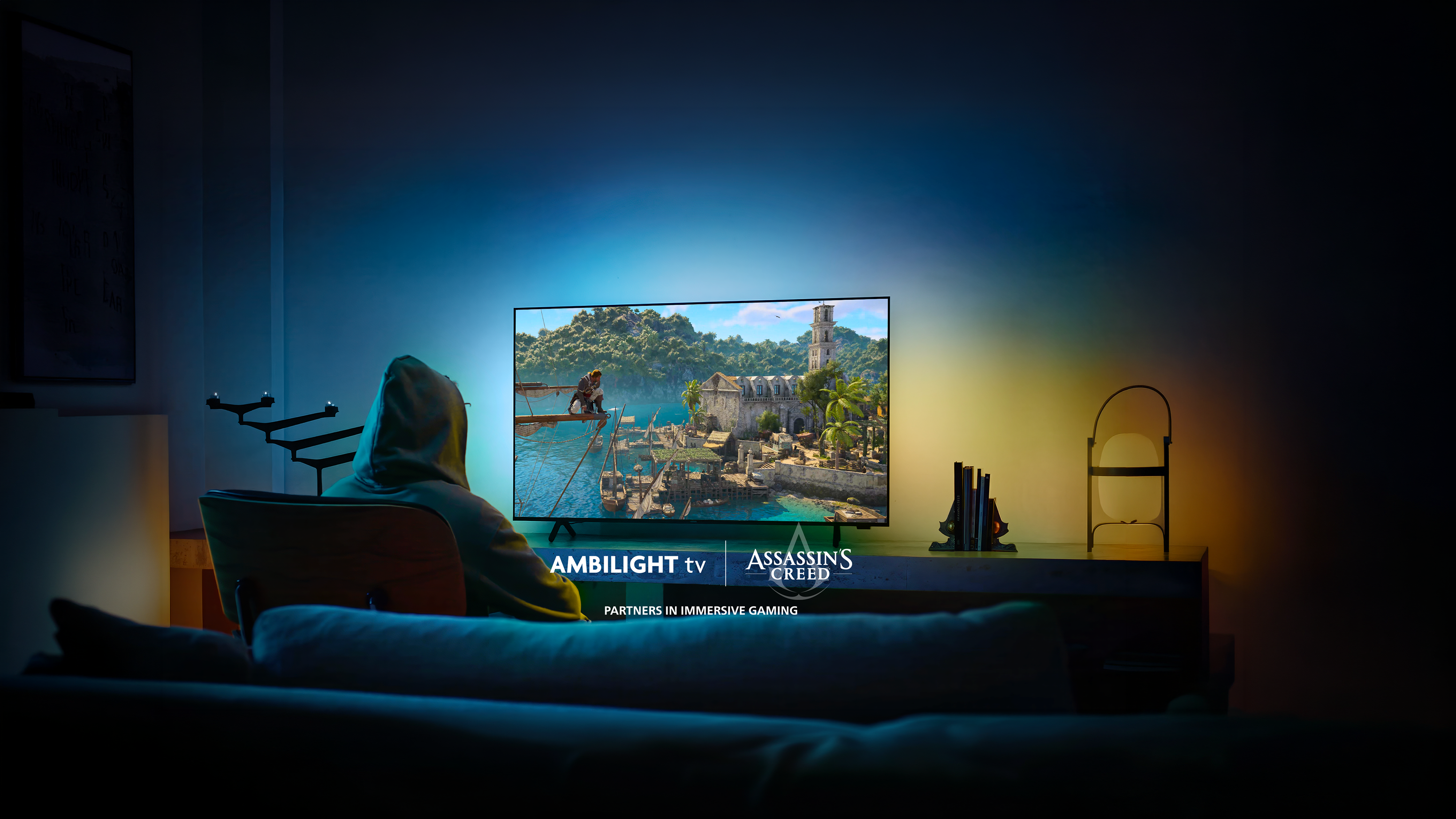Persoon in een hoodie speelt Assassin’s Creed op een Philips Ambilight TV in een donkere kamer, met blauw en amberkleurig licht op de muur achter het scherm.