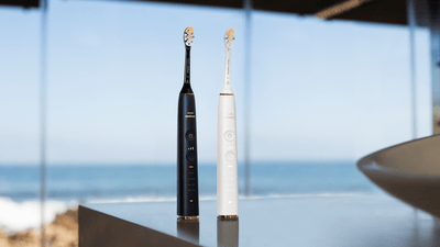 Two Philips electric toothbrushes are stood upright on a counter in a large room with a view of the ocean in the background.