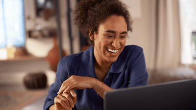 A young woman with dark, curly hair is looking at a laptop screen with a big smile. A living room is in the background.