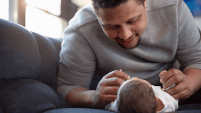 A man attends to his young infant lying on a blue sofa, carefully cleaning the milk coming out of his baby's nose.