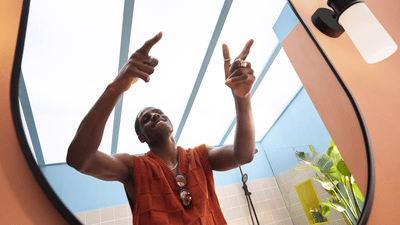 A young Black man in a sleeveless orange top is posing in front of a mirror in a bathroom.