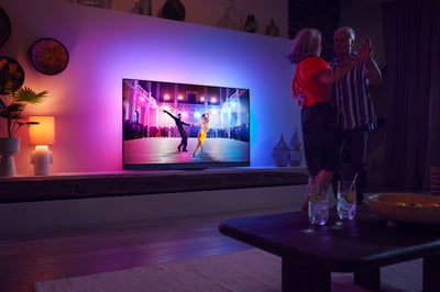 Paar tanzt im Wohnzimmer und schaut einen Philips Ambilight Fernseher mit farbigem Ambient-Licht.