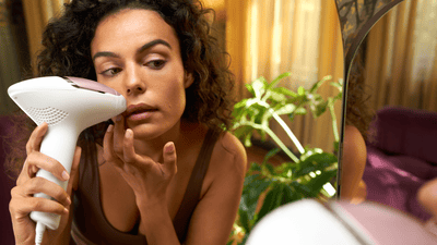 A young woman with dark curly hair leans towards a mirror to remove facial hair with a pink Philips Lumea device