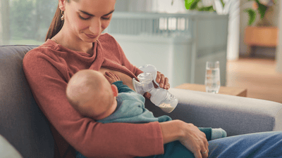 A woman breastfeeds her young infant while simultaneously expressing milk using a Philips Avent breast pump on a sofa.