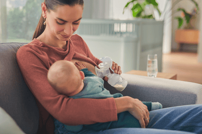 A woman breastfeeds her young infant while simultaneously expressing milk using a Philips Avent breast pump on a sofa.