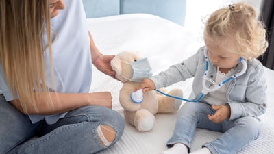 Copil care se joacă de-a medicul cu un ursuleț, alături de mamă