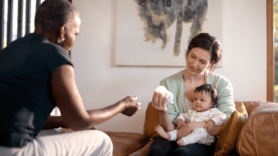 A woman sits on a brown sofa, holding her infant and a Philips Avent baby bottle. A man sits across from them.