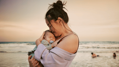 A woman cradles her sleeping baby while standing on an occupied beach. The baby is using a Philips Avent soother.