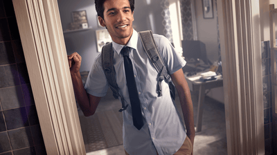 A smartly dressed young man smiles as he leans on the edge of a doorframe in a brightly lit house.