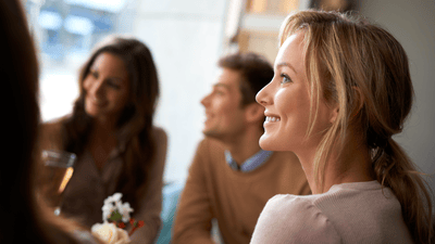 A blonde woman smiles as she engages in conversation with a group of adults holding glasses of alcohol.