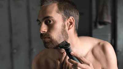 A shirtless man shaves his beard to match his round face using a Philips OneBlade.