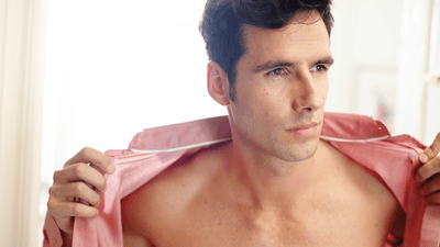 A man with dark hair wearing an unbuttoned pink shirt is looking to the side of the image frame.