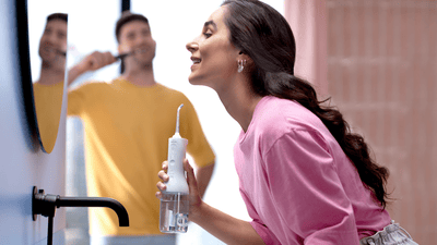 A woman is looking at her teeth in the mirror after using a Philips Air Flosser. A man in the background is brushing his teeth.