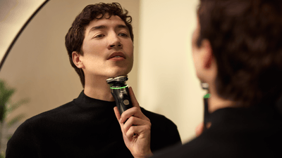 A man wearing a black turtle neck jumper shaves his beard using a black Philips shaver in front of a mirror.