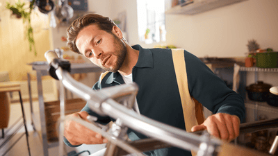 A man with a stubble beard wearing a dark green shirt over a white tank top is adjusting a bicycle.