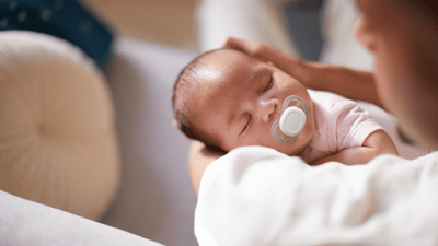 A woman soothes her sleeping infant with a Philips Avent Soother while sitting on a cream sofa.