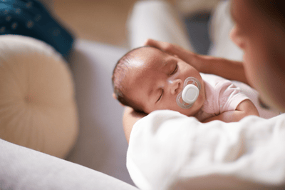 A woman soothes her sleeping infant with a Philips Avent Soother while sitting on a cream sofa.