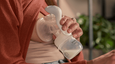 A blonde woman wearing a beige sweater sits in a boho-themed lounge while using a manual breast pump.
